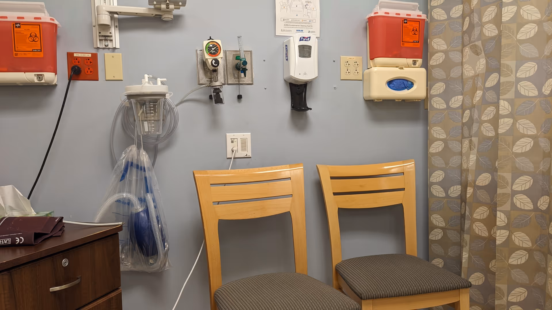 Two wooden chairs sit beside medical equipment mounted on a clinic wall.