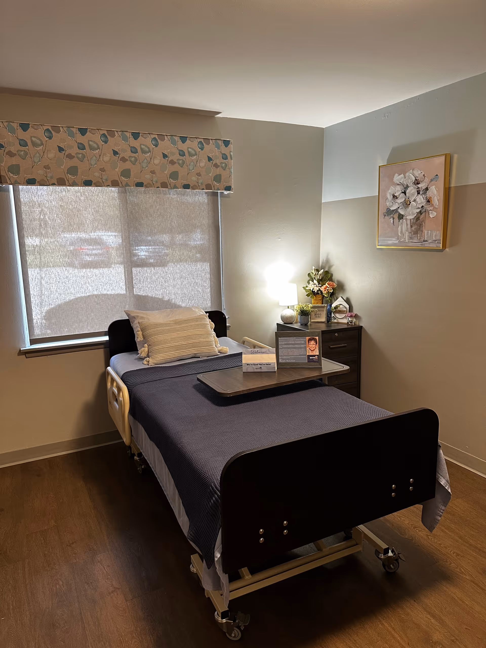 A single hospital-style bed with a dark blue blanket and a white pillow in a room with wooden flooring. There is a window with a floral valance and a roller shade partially down. Next to the bed is a small dark wooden nightstand with a lamp, flowers, and framed pictures. A painting of white flowers in a vase hangs on the wall above the nightstand.