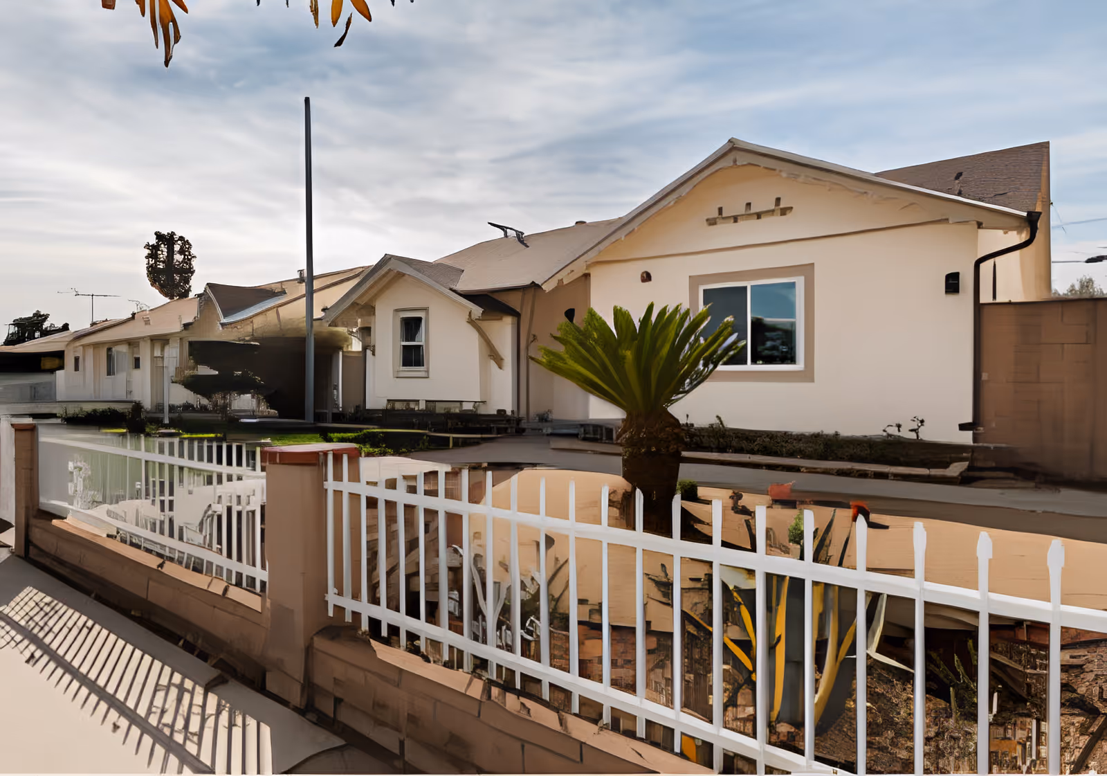 Front exterior of a single-story residential building with a white metal fence and a small palm in the yard.