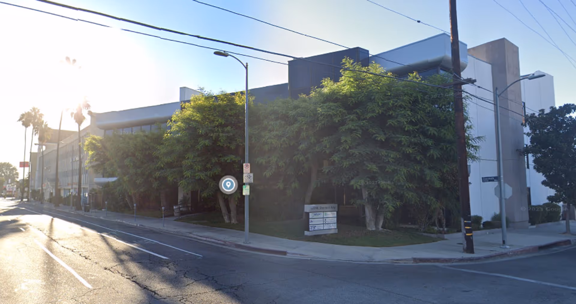 Street view of a modern facility with a tree-lined sidewalk on a sunny morning.
