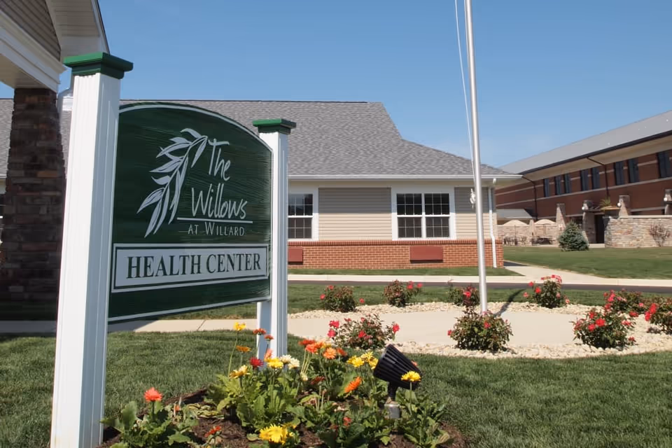 A white-and-green sign reading "The Willows at Willard Health Center" stands in a flowered lawn in front of a single-story brick and siding building.
