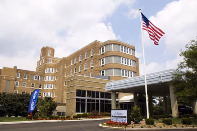 Multi-story brick senior living building with a covered entrance, American flag, landscaping, and a sign for The Villages at Historic Silvercrest.