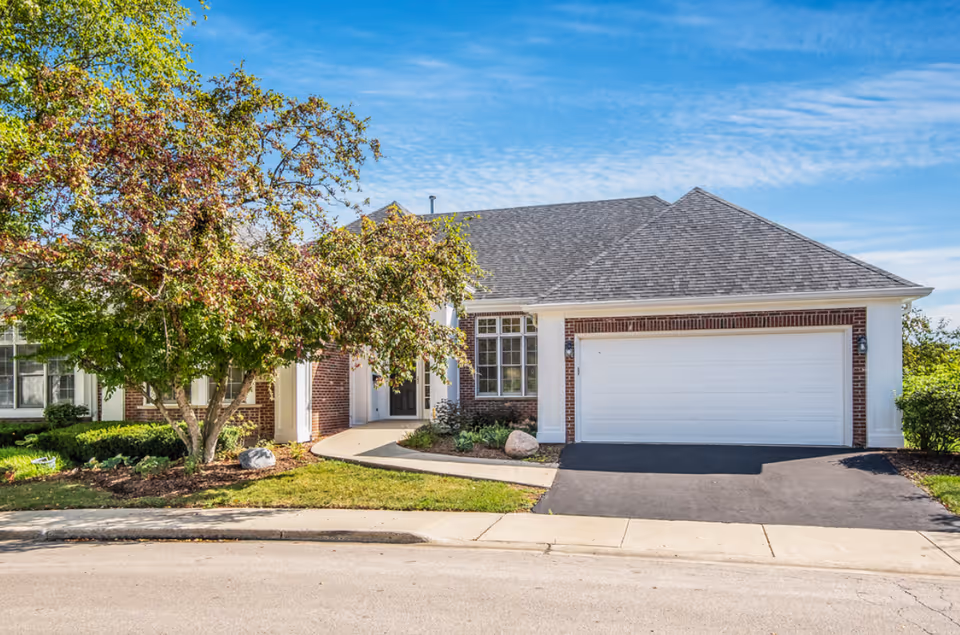 Exterior view of a single-story brick and white siding house with a two-car garage, a paved driveway, a curved walkway leading to the front door, and a tree with green and reddish leaves in the front yard under a blue sky.