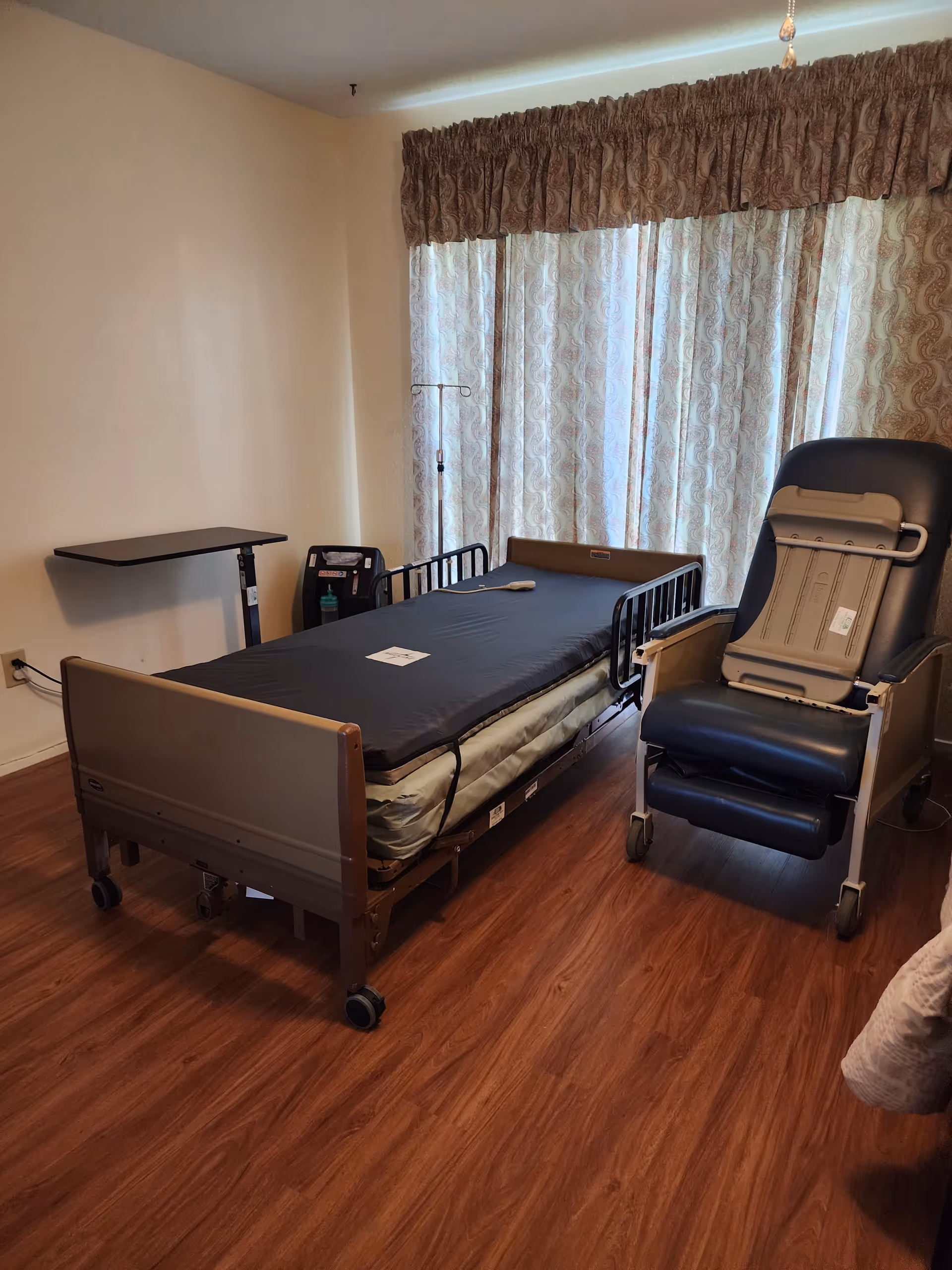 Hospital-style patient bed with rails, IV stand, and recliner chair in a curtained hospice room.