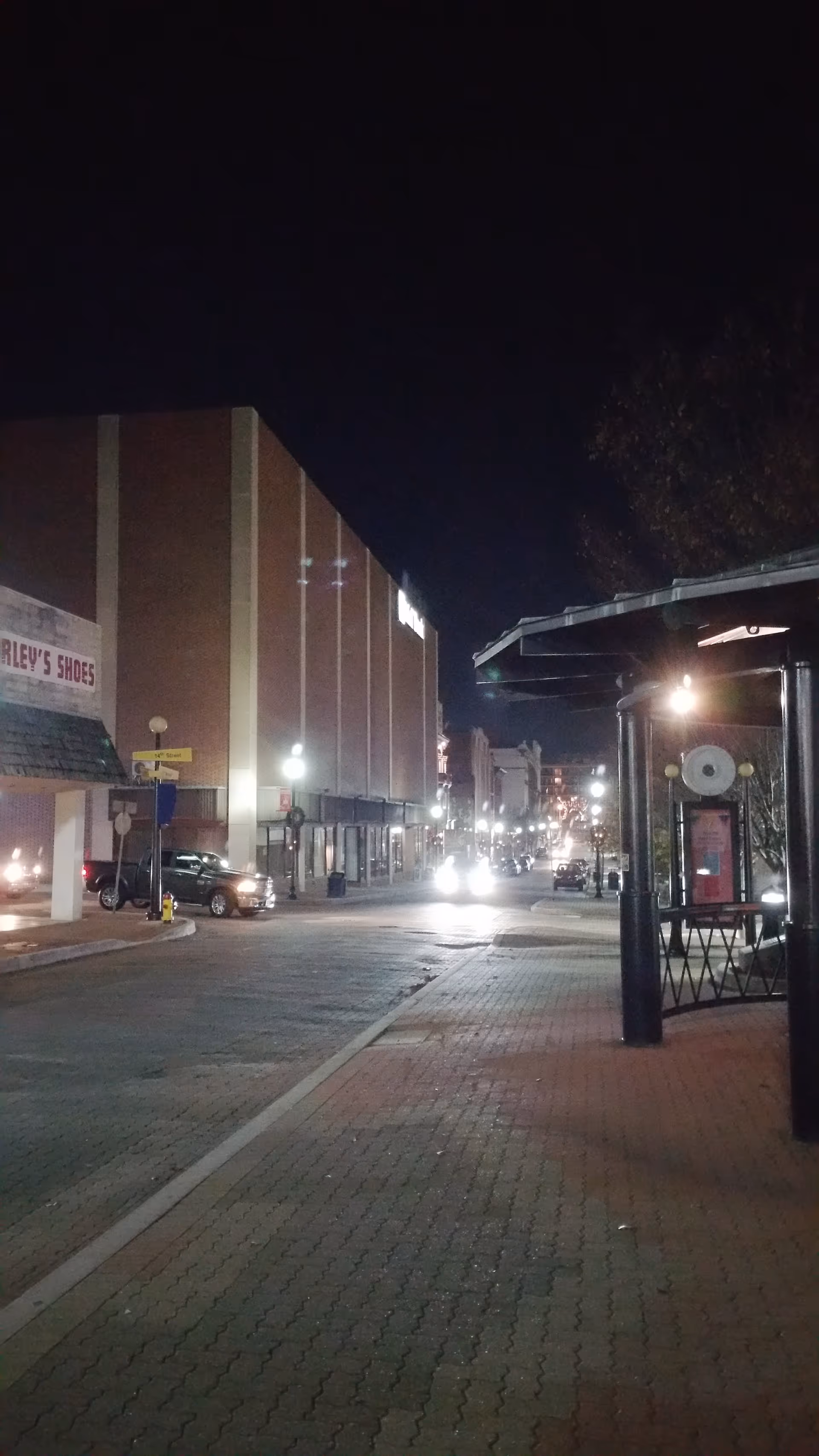 Nighttime street scene with brick buildings, storefronts, and a lit sidewalk.