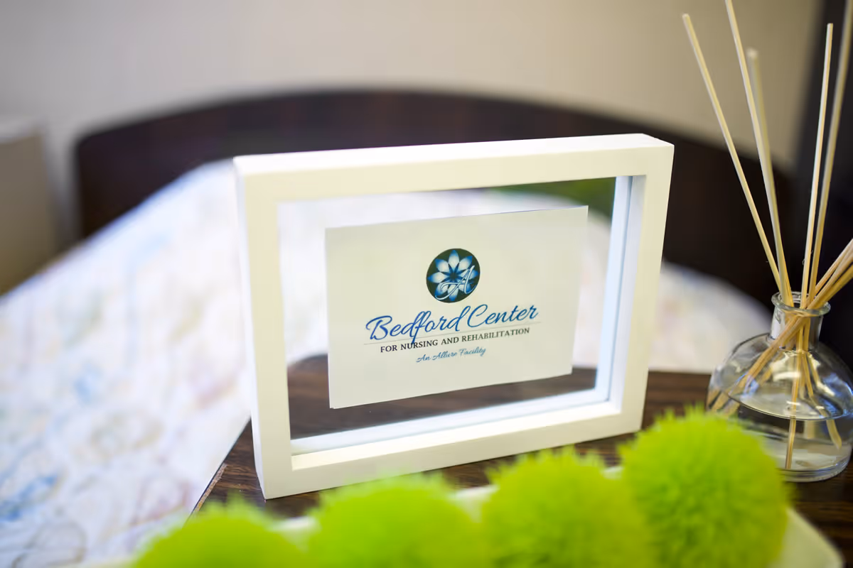 A close-up of a white picture frame on a wooden surface displaying the Bedford Center for Nursing and Rehabilitation logo. In the background, there is a blurred bed with a patterned blanket. To the right of the frame, there is a glass container with reed diffuser sticks. Green decorative pom-poms are visible in the foreground.