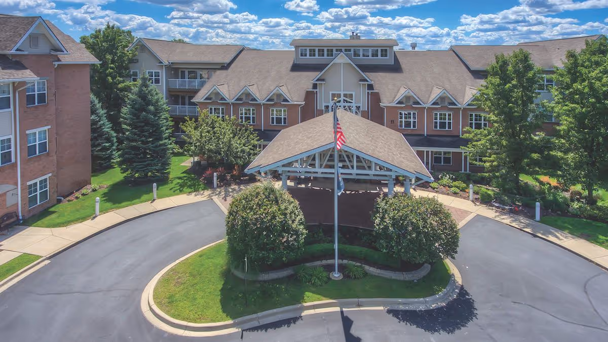 A large brick senior living building with a covered entrance, circular driveway, and flag