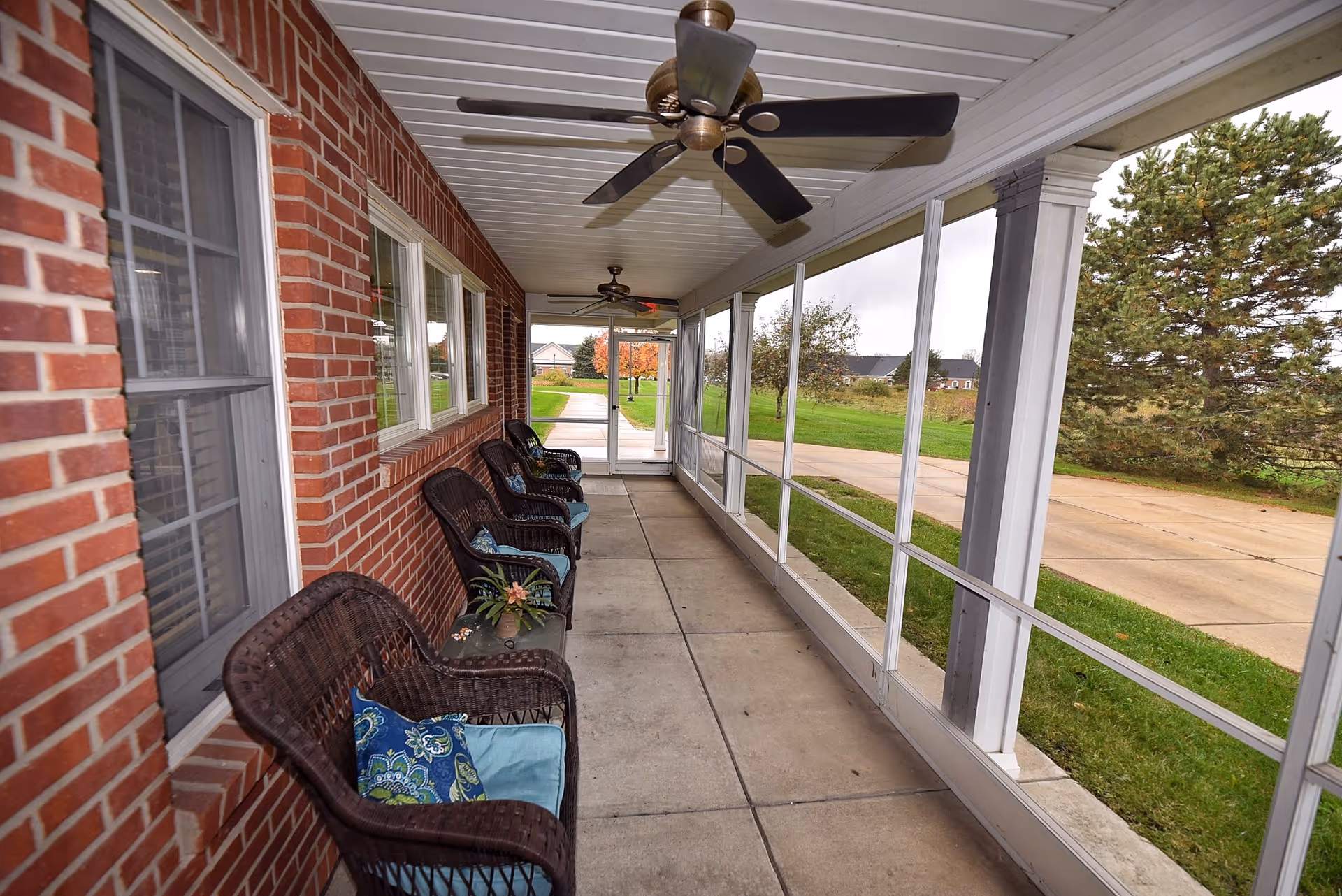 Covered brick porch with wicker chairs, ceiling fans, and large windows along a walkway.