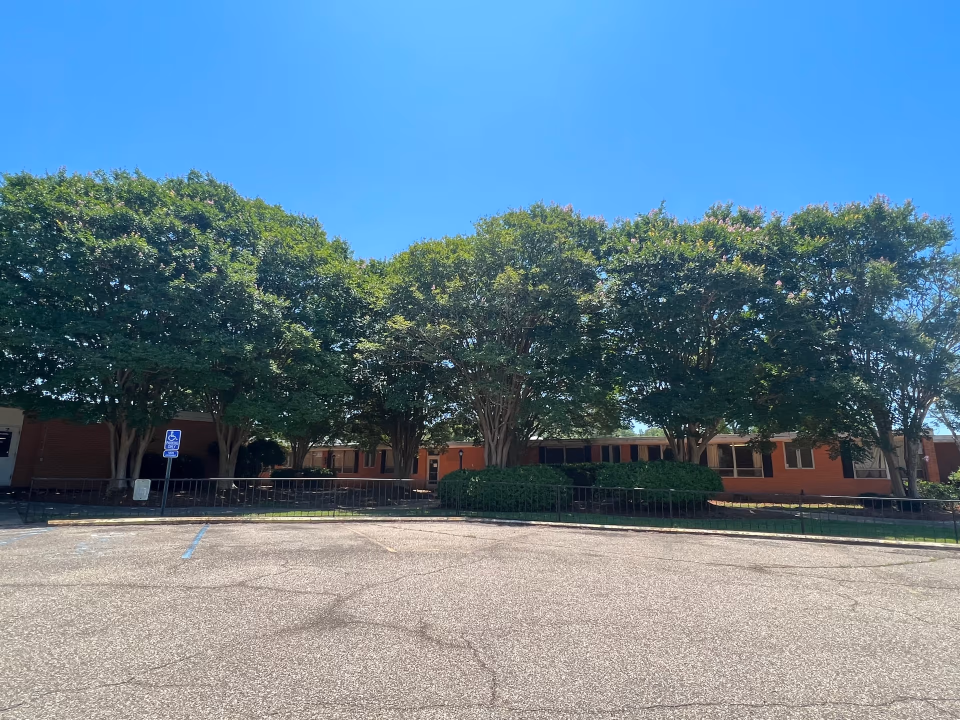 Exterior view of Leslie Lakes Retirement Center showing a single-story brick building partially obscured by large green trees under a clear blue sky. In the foreground is an empty parking lot with a handicapped parking sign visible.