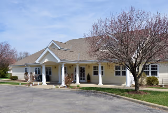 Exterior view of a single-story building with beige siding and a gabled roof. The entrance features white columns supporting a small porch roof. There are leafless trees and some shrubs around the building, with a paved driveway in front.