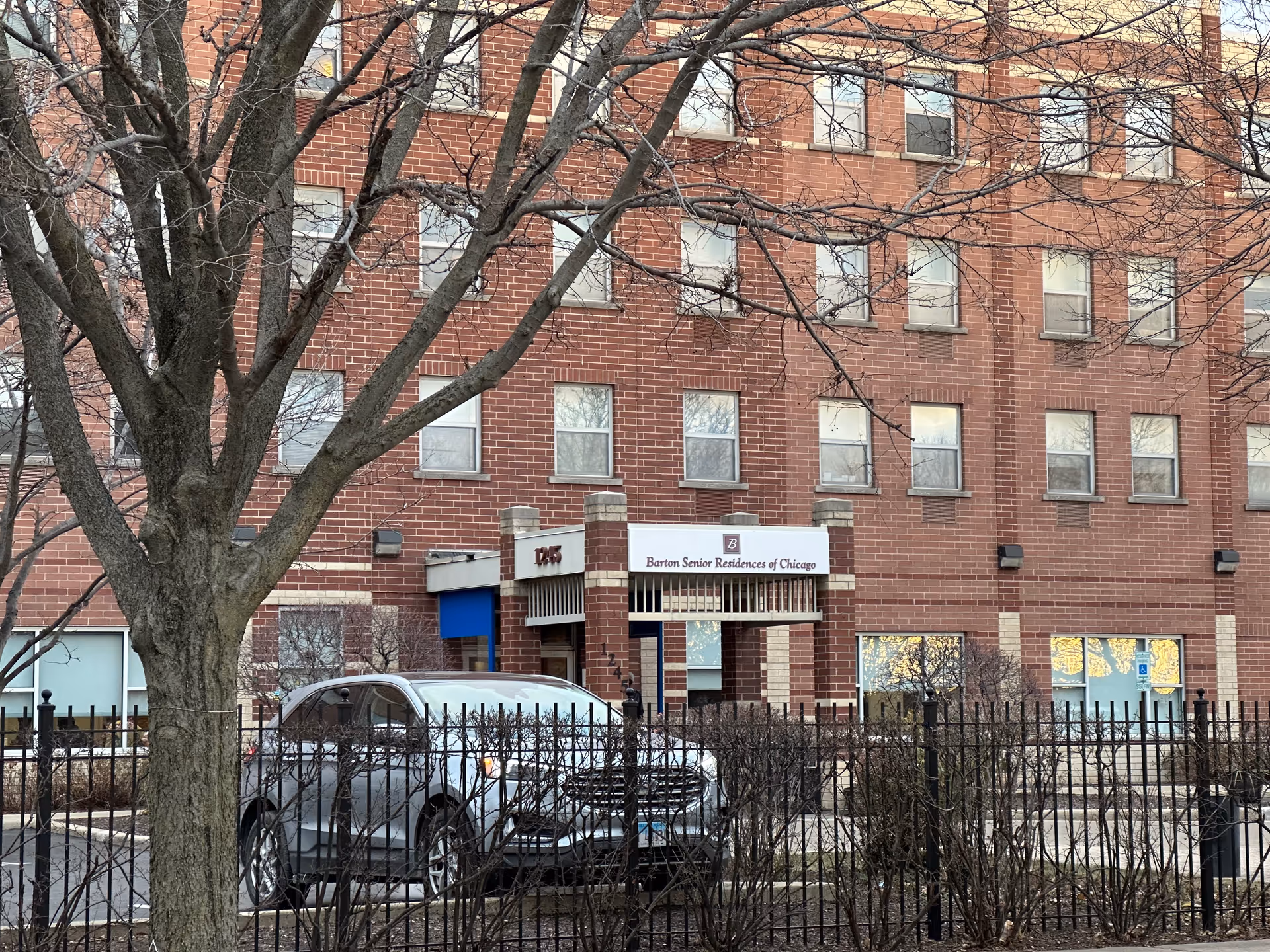 Front facade of a red-brick Barton Senior Residences building with its entrance canopy, a parked car, and a leafless tree in the foreground.