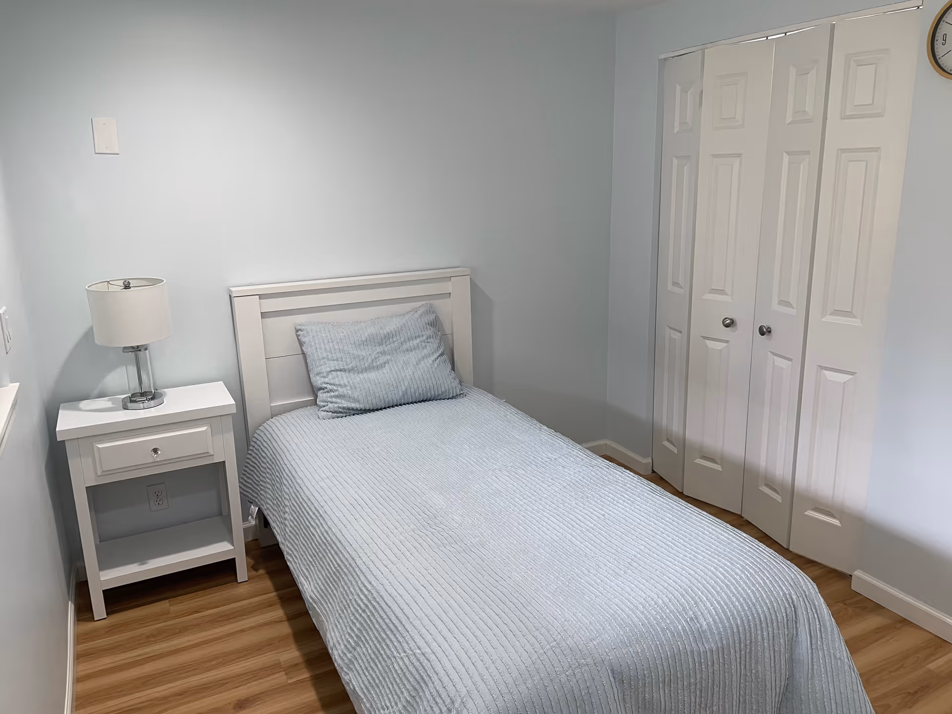 A small bedroom with a single bed covered in a light blue textured bedspread and matching pillow. Next to the bed is a white nightstand with a drawer and a modern lamp on top. The room has light blue walls, a wooden floor, and a white closet with double doors. A round clock is partially visible on the wall above the closet.