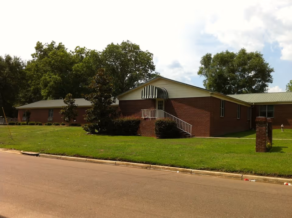 Exterior view of a single-story brick building with a green lawn and trees surrounding it. The building has a small staircase leading to a door with a striped awning above it. The sky is partly cloudy.