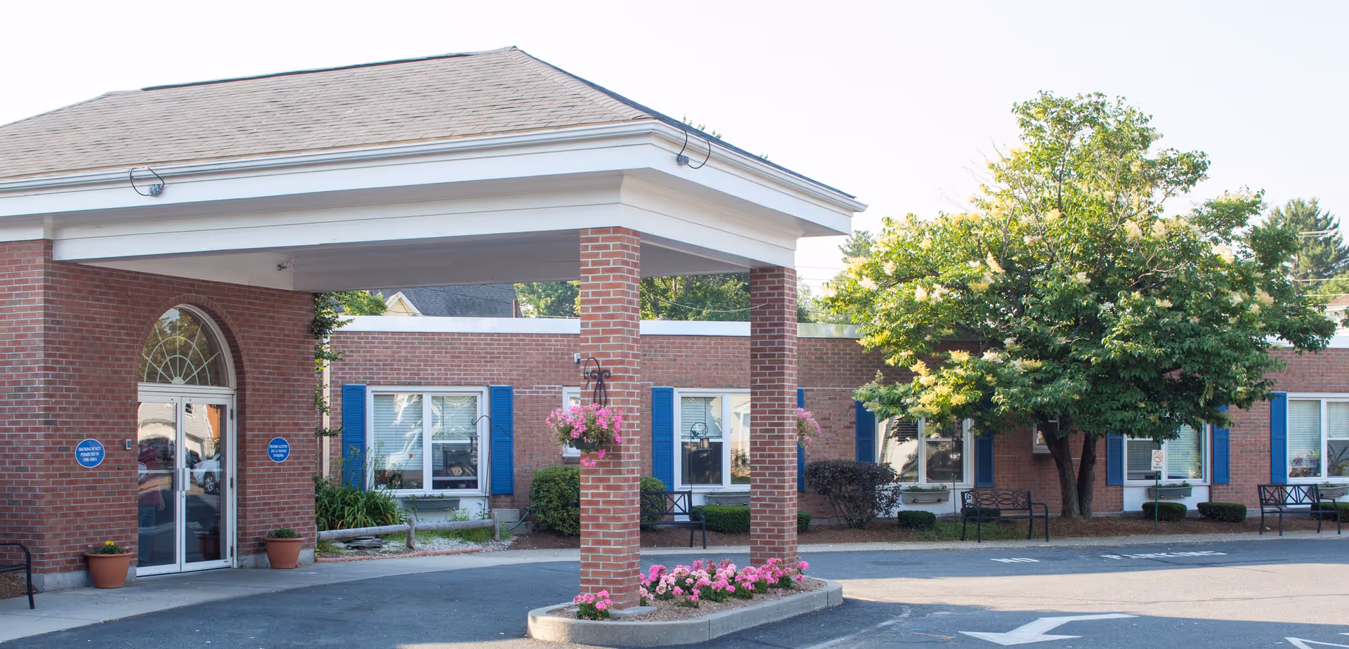 Exterior view of a single-story brick building with a covered entrance supported by brick columns. The building has several windows with blue shutters and is surrounded by landscaping including potted plants, flower beds, and a large tree. There are benches near the windows and a paved driveway in front.