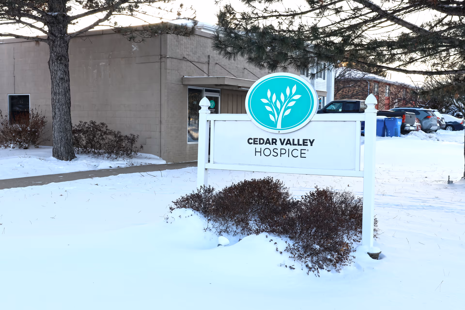 Winter scene outside Cedar Valley Hospice with a sign, snow-covered ground and shrubs.