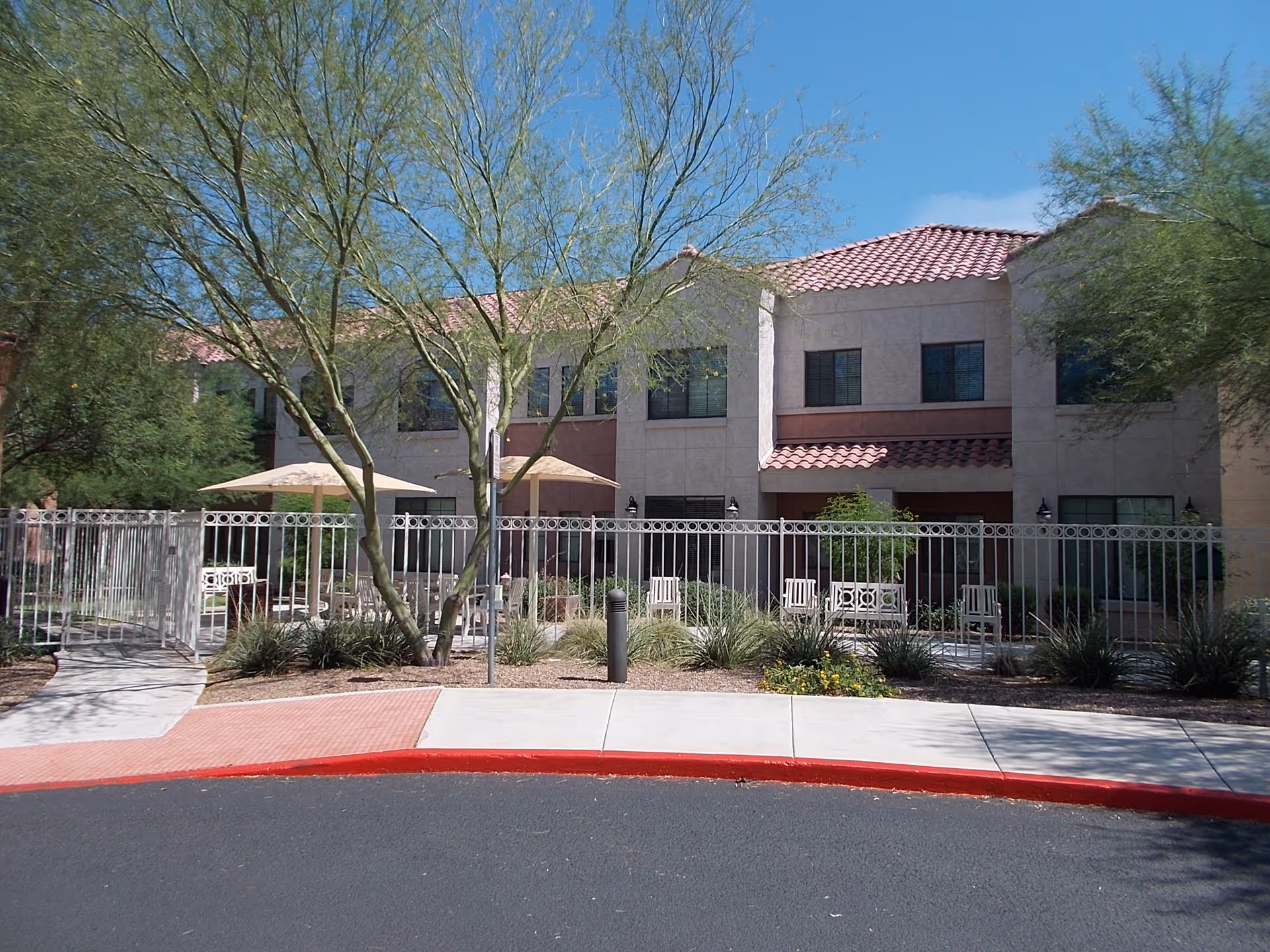 Front exterior of a two-story building with a fenced patio area, umbrellas, benches, and desert landscaping.