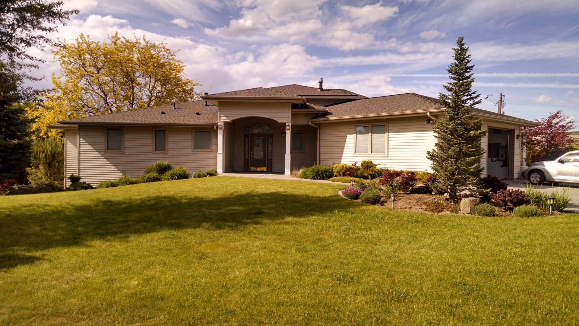 Single-story beige residential facility with front entrance, well-kept lawn, and surrounding shrubs.