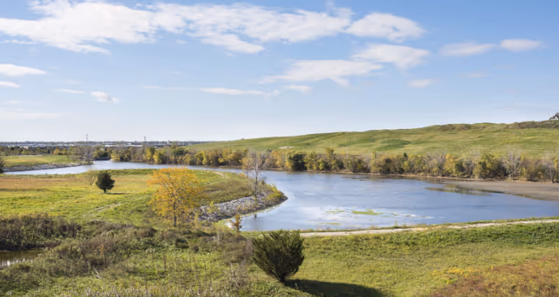 A scenic outdoor view of a winding river surrounded by green grassy fields and trees under a partly cloudy blue sky.