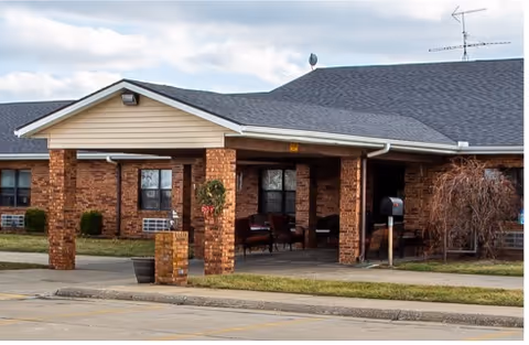 Exterior view of a brick building with a covered entrance supported by brick columns. There are chairs and a mailbox near the entrance, and the building has a dark shingled roof with an antenna on top. The sky is partly cloudy.