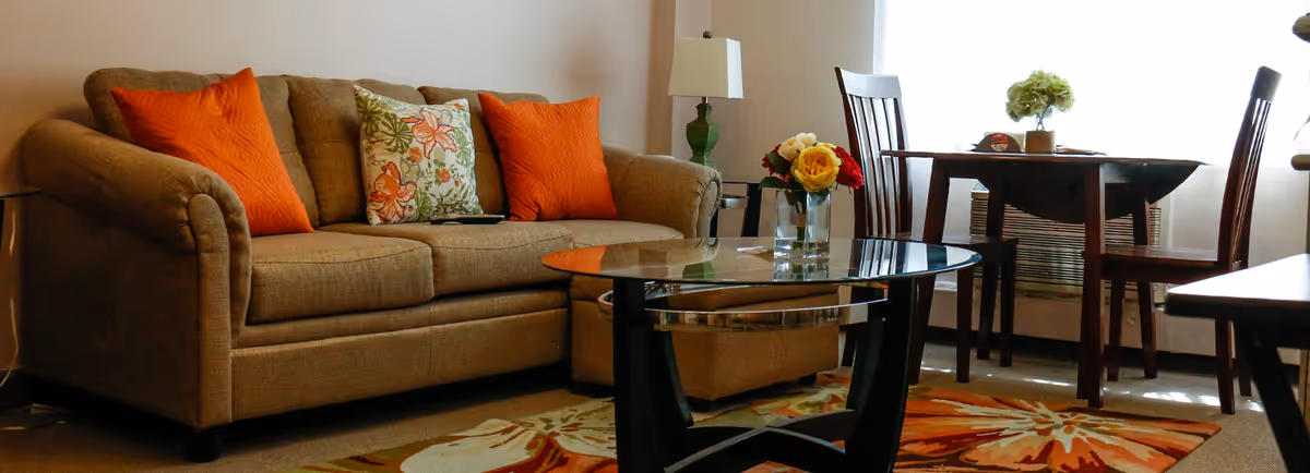 A cozy living room with a tan sofa accented by orange and floral pillows, a glass coffee table, and a small dining table by a window.