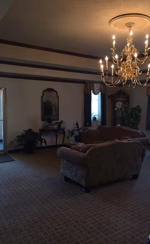 Dimly lit senior living room with grand chandelier, sofas, and potted plants.