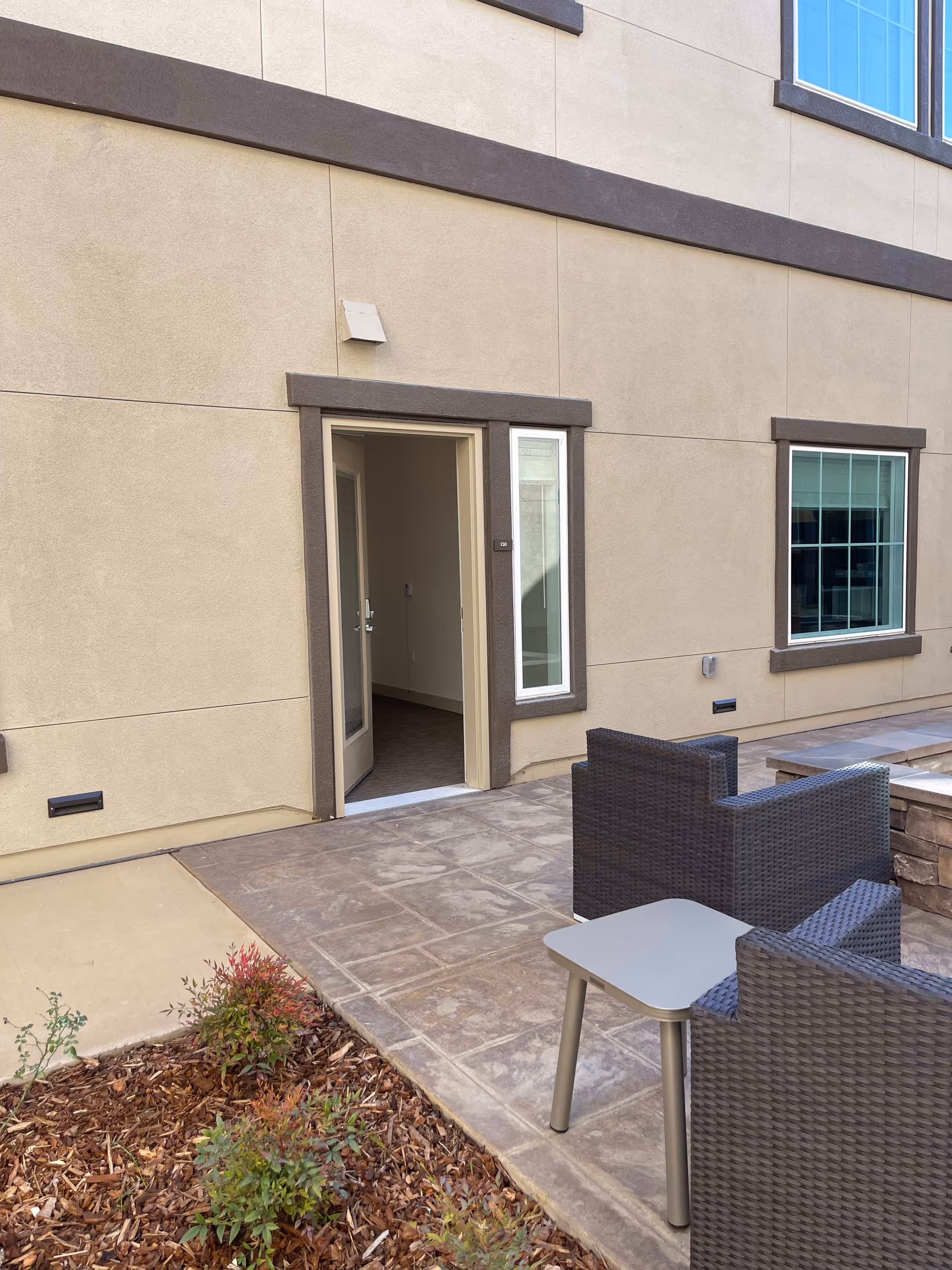 Outdoor patio area with wicker chairs and a small table in front of an open doorway to a beige building and adjacent landscaping.