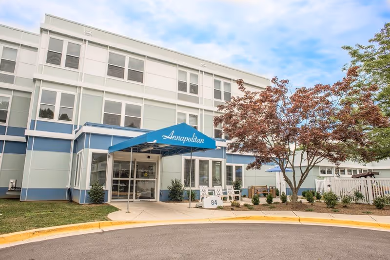 Exterior view of Annapolitan Assisted Living facility showing the main entrance with a blue awning labeled 'Annapolitan'. There are trees and shrubs around the entrance, a small seating area with chairs, and a curved driveway in front.