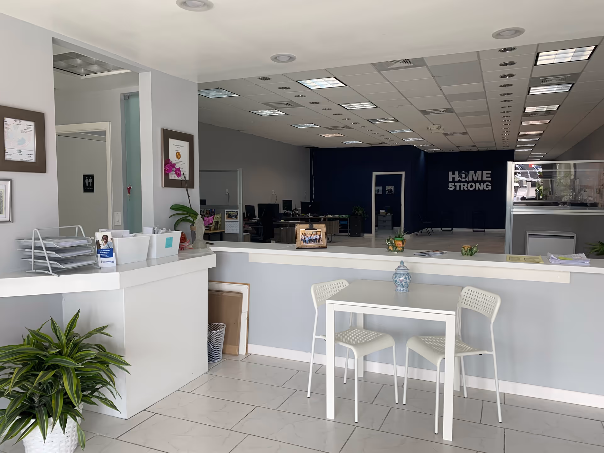Reception area with a white counter, small table and chairs, plants, and office space.