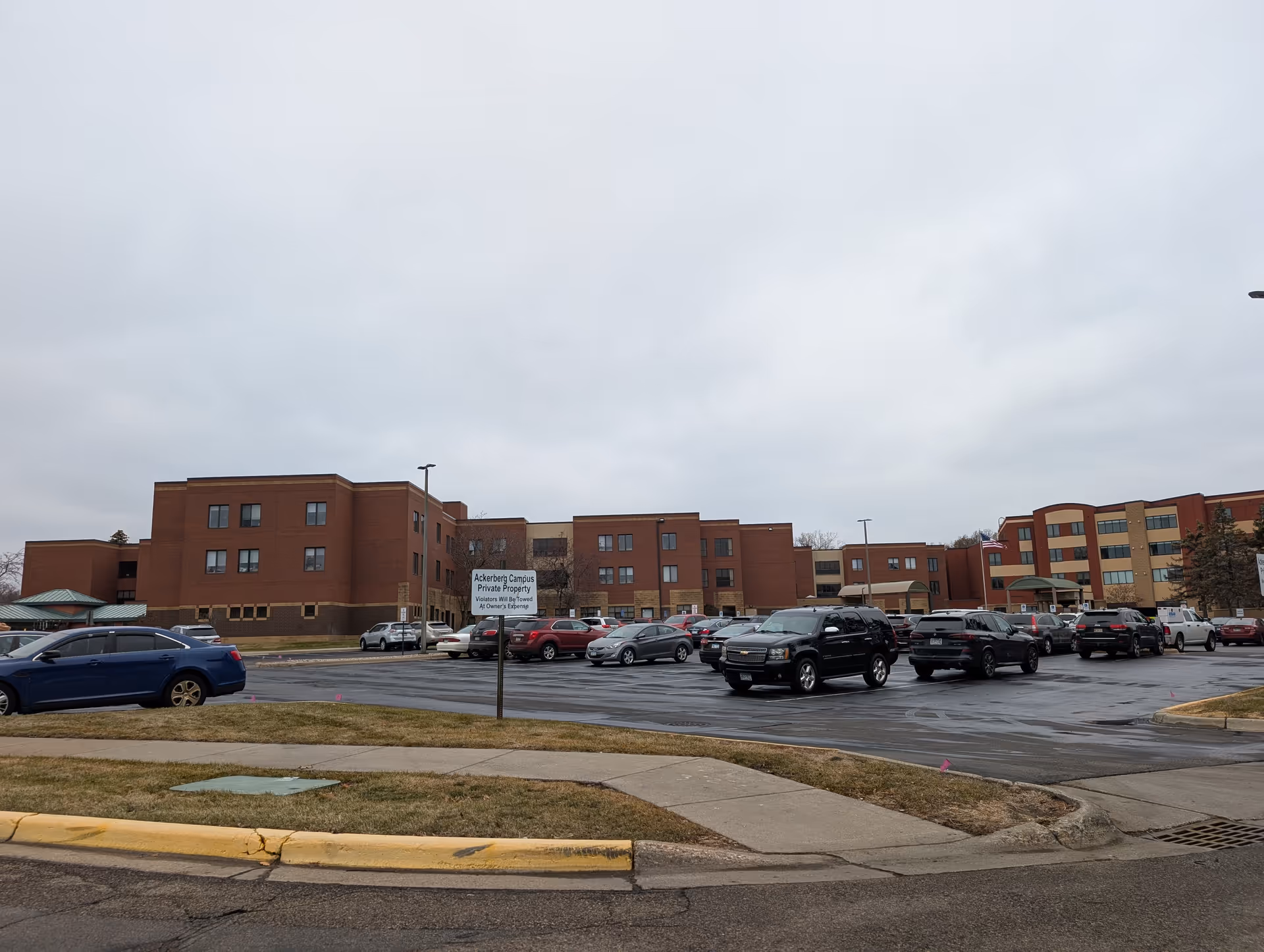 Exterior view of a large brick building complex with multiple floors, a parking lot filled with cars in front, and a cloudy sky overhead. A sign in the parking lot reads 'Ackerberg Campus Private Property Violators Will Be Towed At Owner's Expense.'
