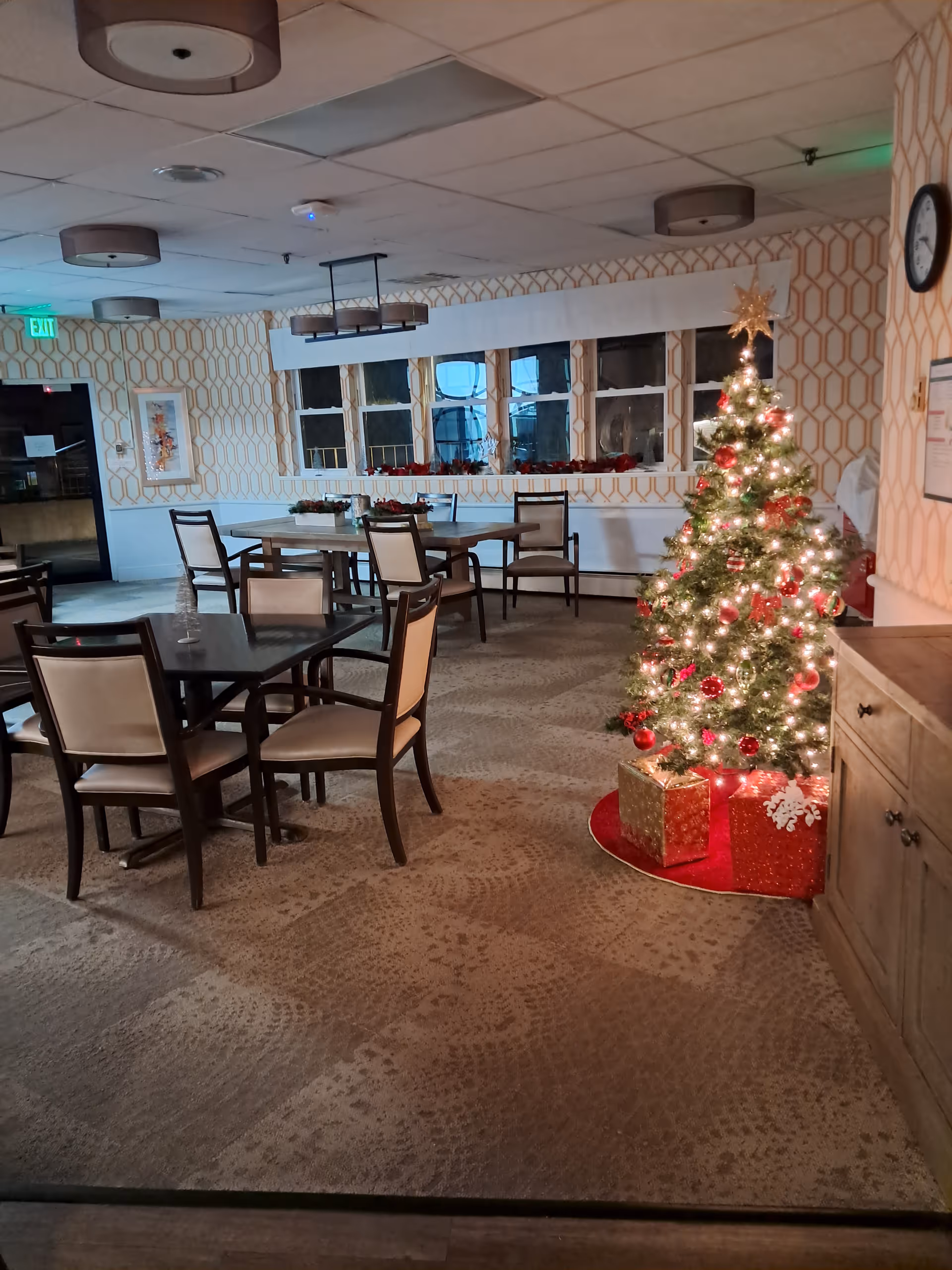 A dining area with tables and chairs, a holiday tree, and wrapped presents nearby.