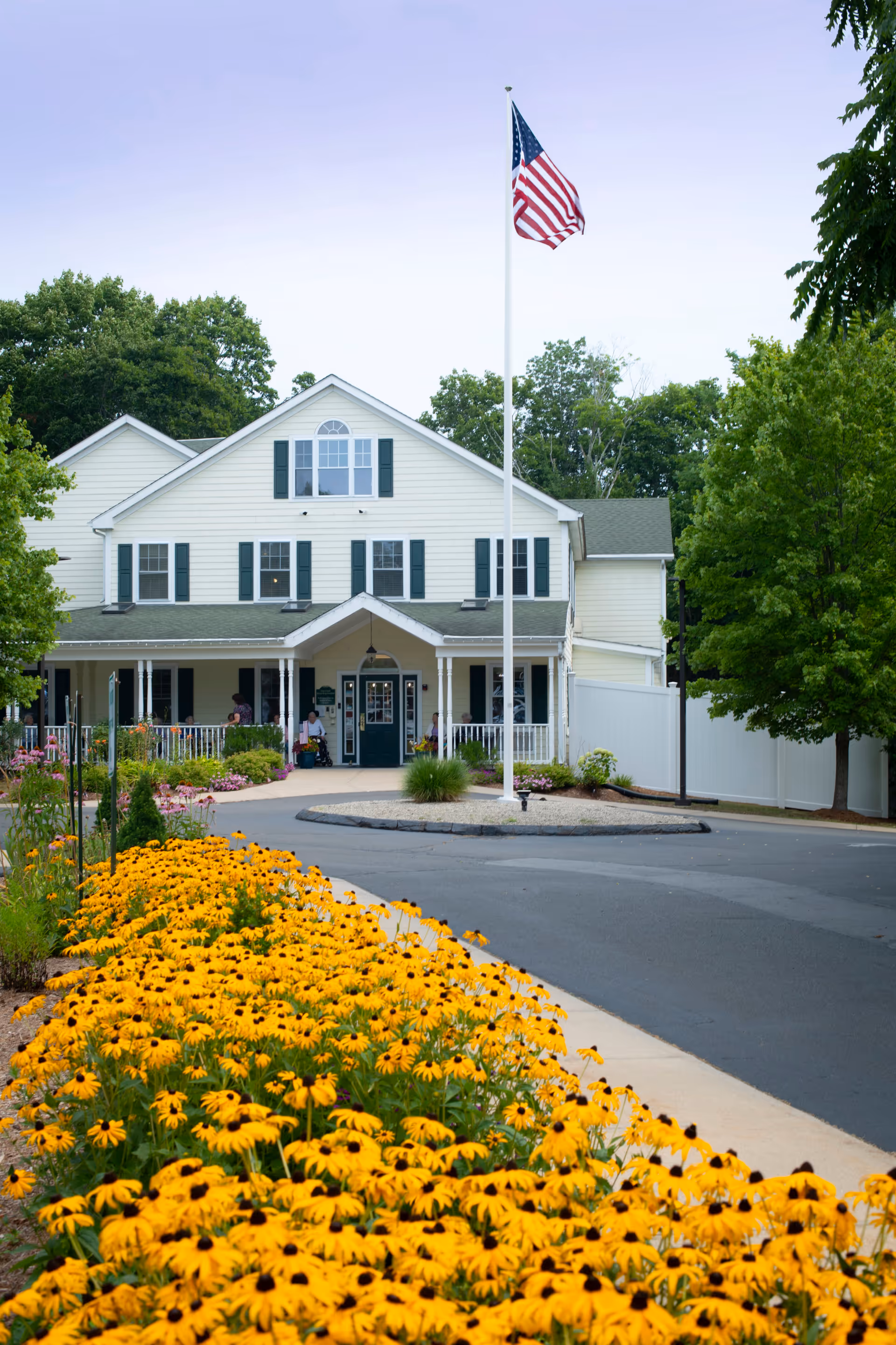 Front exterior view of a two-story white building with green shutters, a covered porch, and an American flag on a tall flagpole in front. There are yellow flowers along the sidewalk leading to the entrance, and trees surrounding the building.