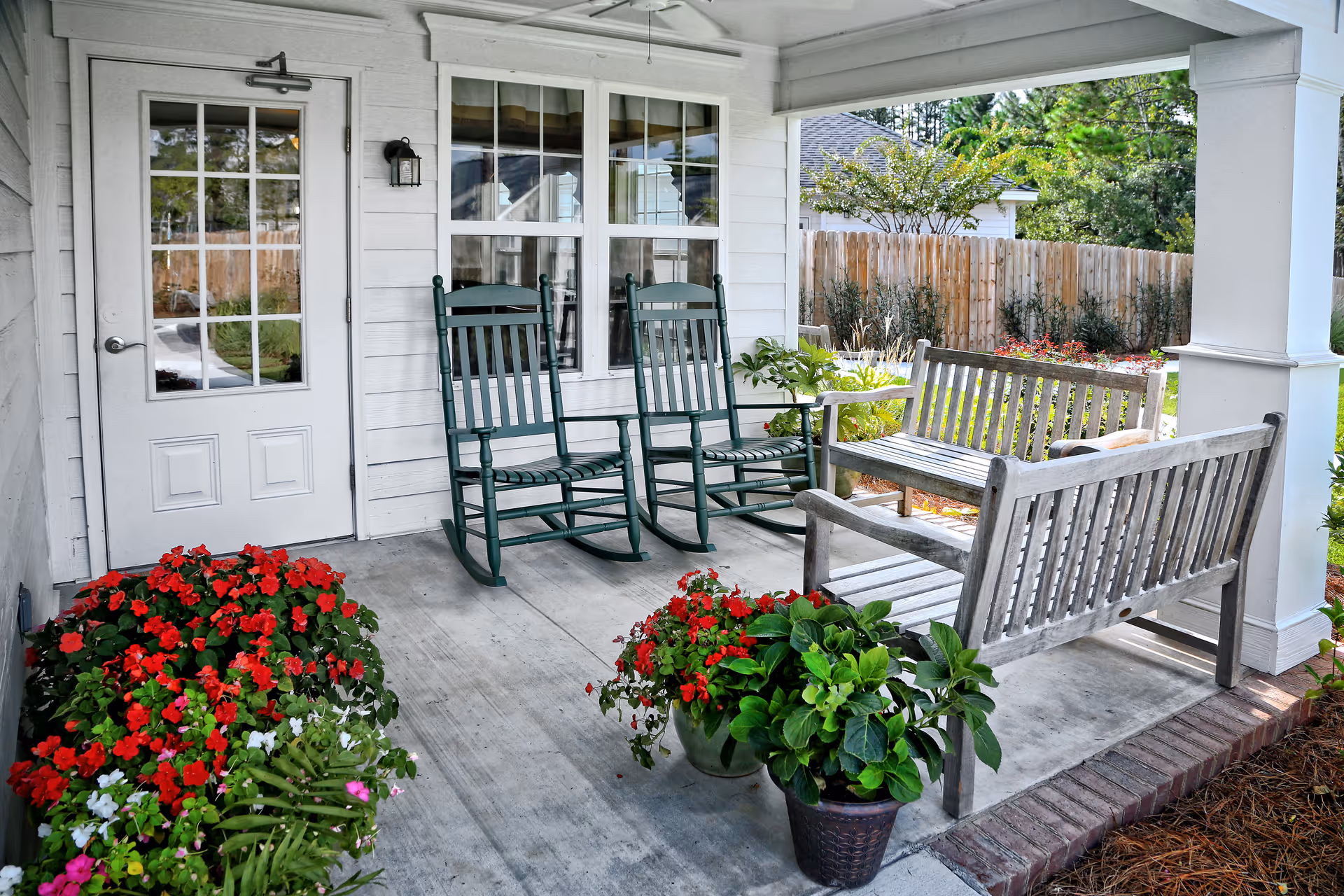 Covered porch with green rocking chairs, wooden benches, and potted red flowers beside a white door and window.