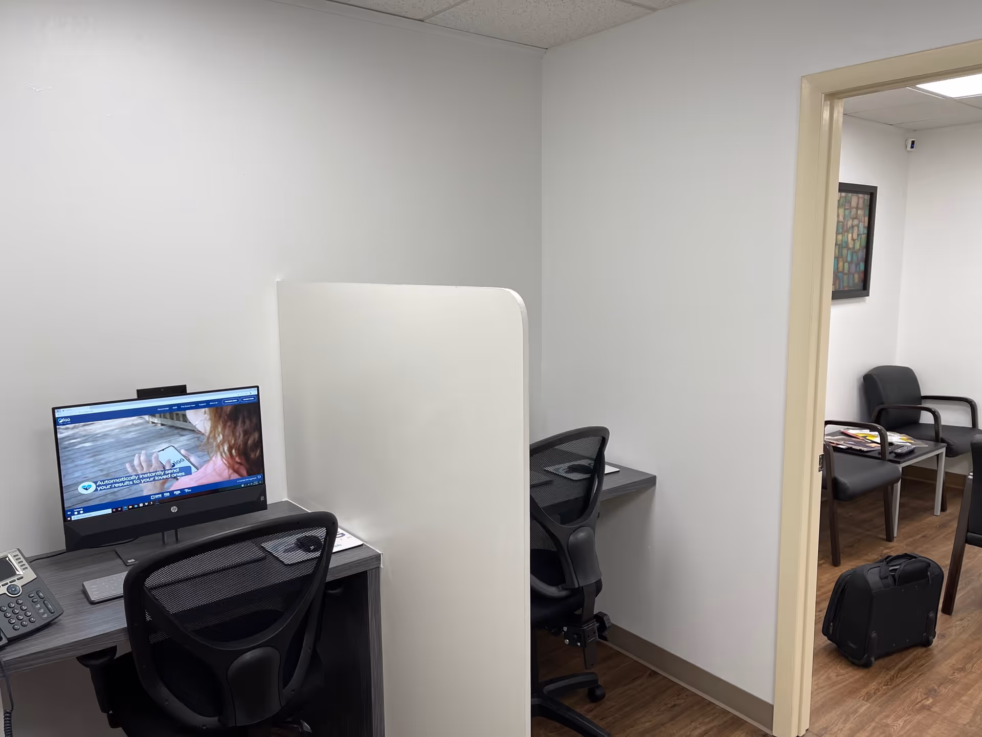 Empty office area with desks, a partition, computer, and a lounge visible through a doorway.