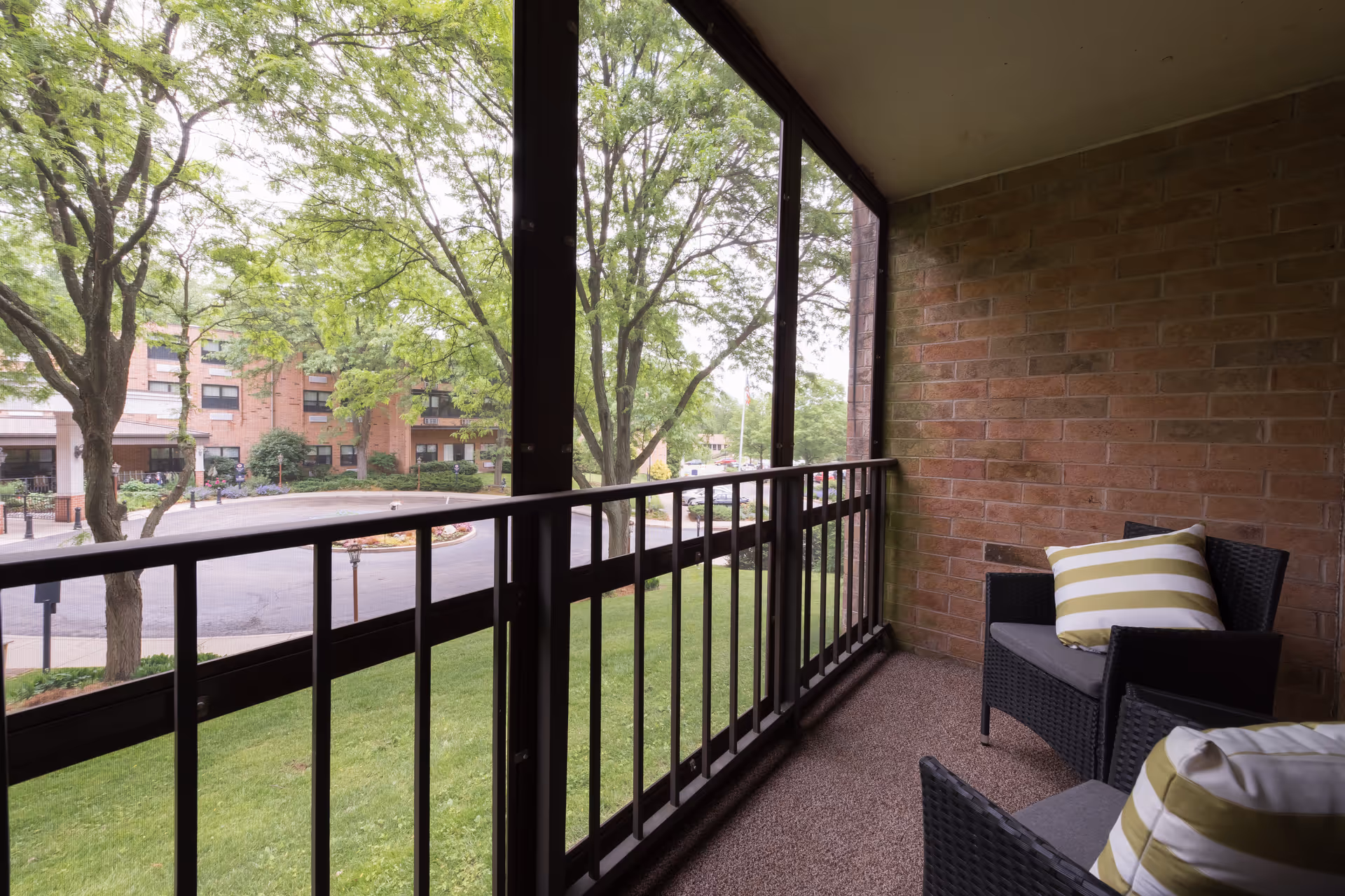 Brick balcony with wicker chairs and striped pillows overlooking trees and a parking lot.