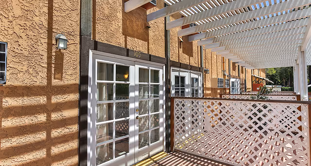 Exterior corridor of The Chateau of Long Beach showing stucco walls, a pergola casting shadows, lattice patio railing, and French doors labeled "105".