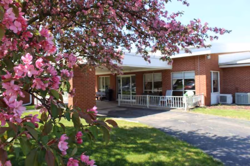 Brick-front entrance of a senior living facility with a covered porch and a pink flowering tree in the foreground.