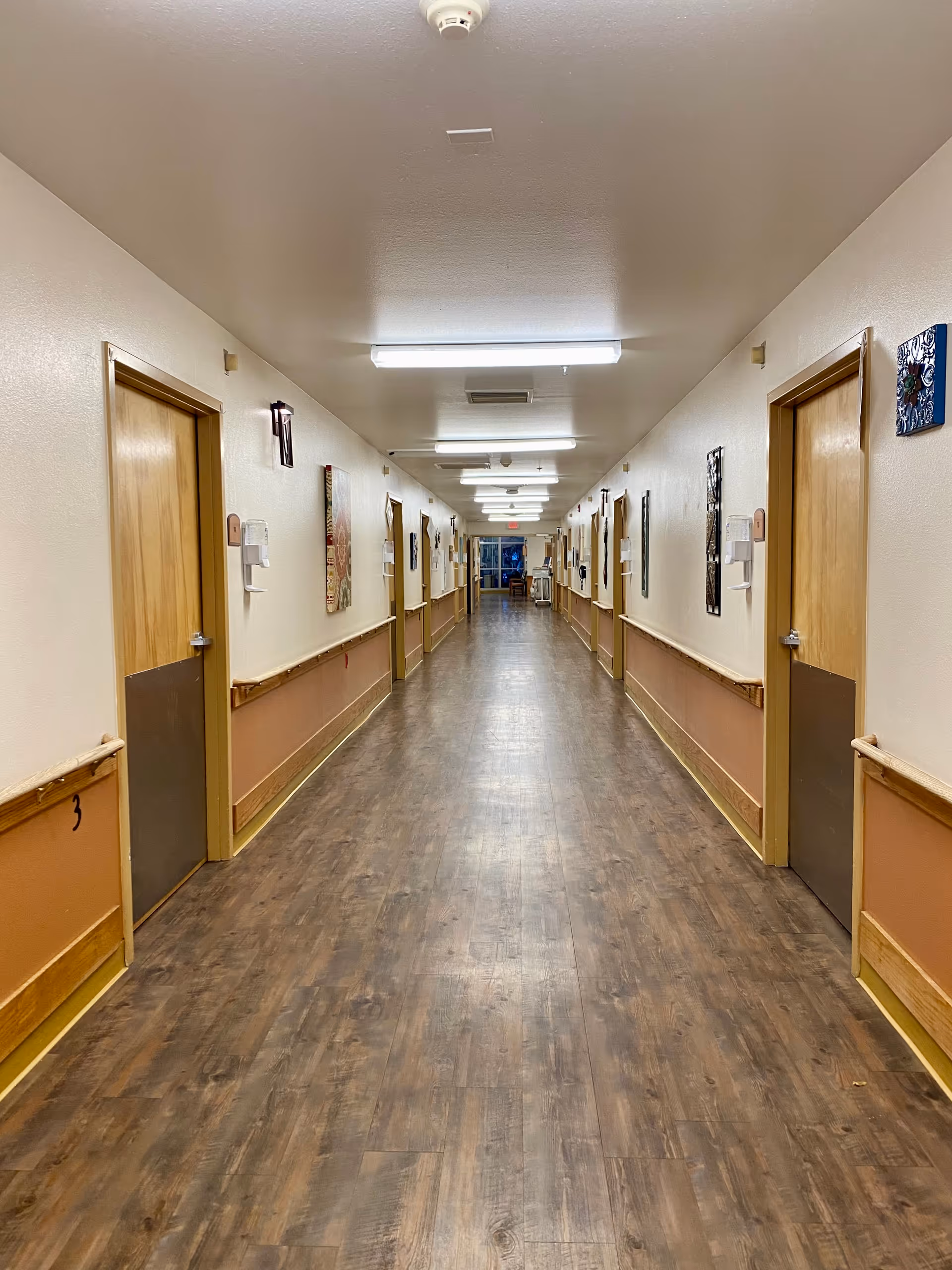 Long institutional hallway with doors on both sides, wooden floor, and bright overhead lights.