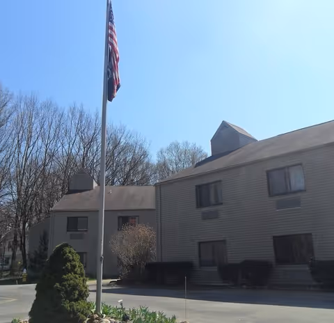 Assisted living facility exterior with flagpole, trees, and blue sky.