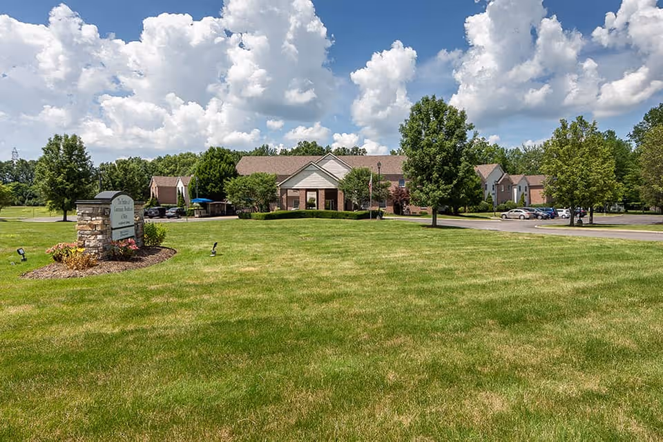 Front view of a low-rise brick senior living facility with a large green lawn, entrance sign, trees, and parked cars under a partly cloudy sky.