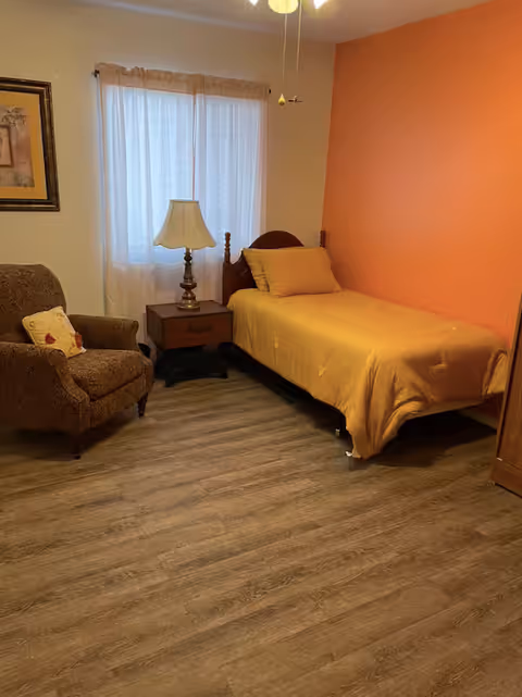 A cozy bedroom with a single bed covered in a yellow bedspread against an orange accent wall. Next to the bed is a wooden nightstand with a lamp. There is a comfortable upholstered armchair with a decorative pillow, a window with sheer curtains, and a framed picture on the wall. The floor is wood with a natural finish.