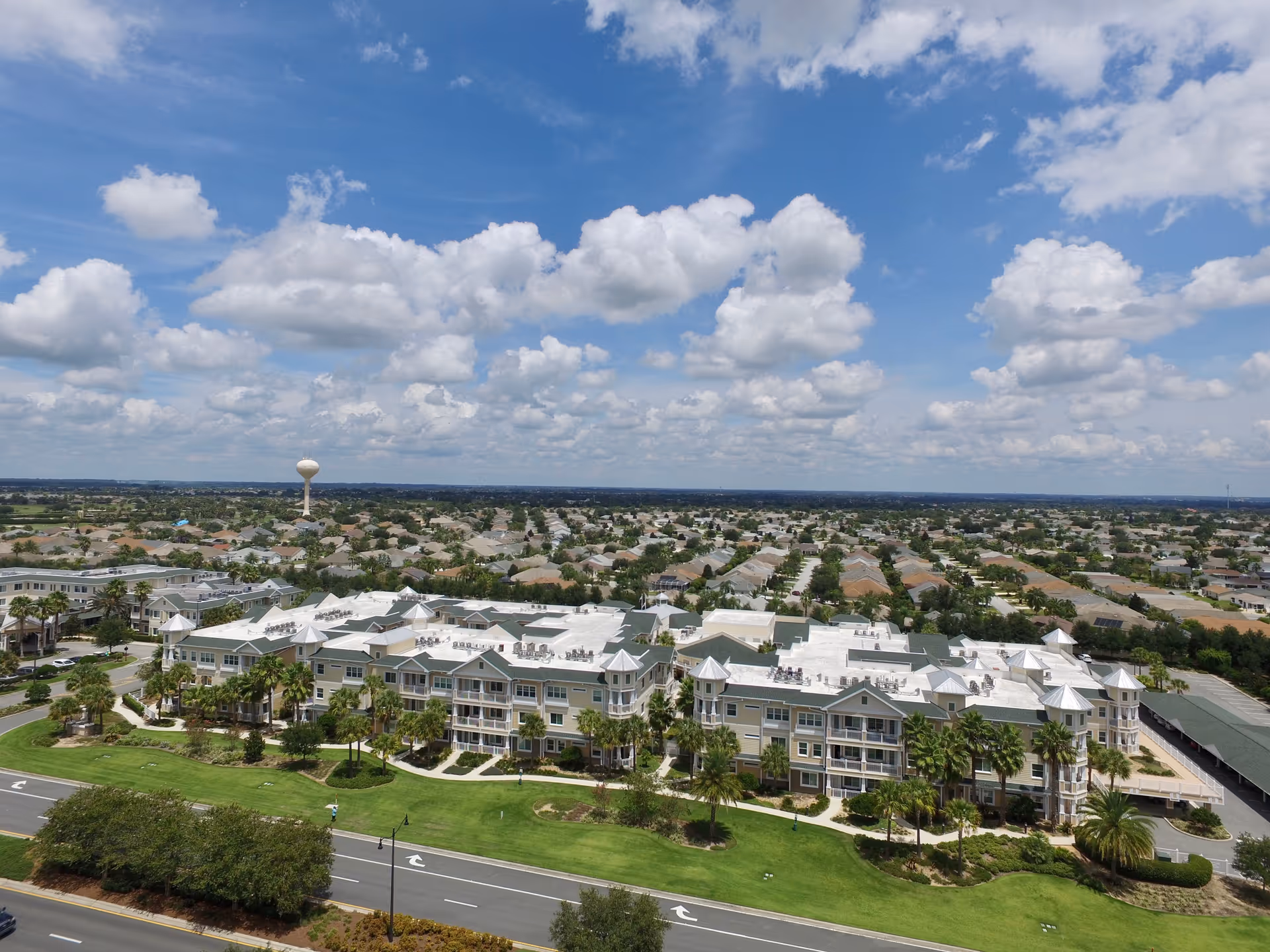 Aerial view of a large senior living facility with multiple connected buildings featuring white roofs and beige walls, surrounded by green lawns and palm trees. In the background, there is a suburban neighborhood with many houses and a water tower under a partly cloudy blue sky.