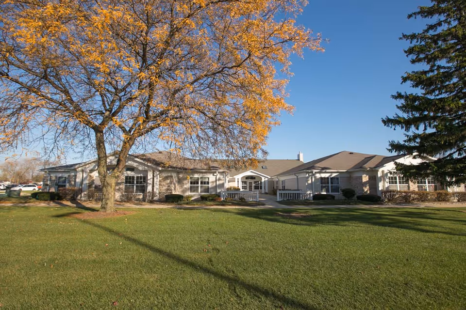 Front exterior of a single-story senior living building with a large lawn and trees showing autumn foliage.