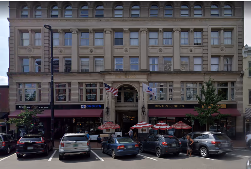 A large historic brick building with storefronts and red umbrellas along a city street.