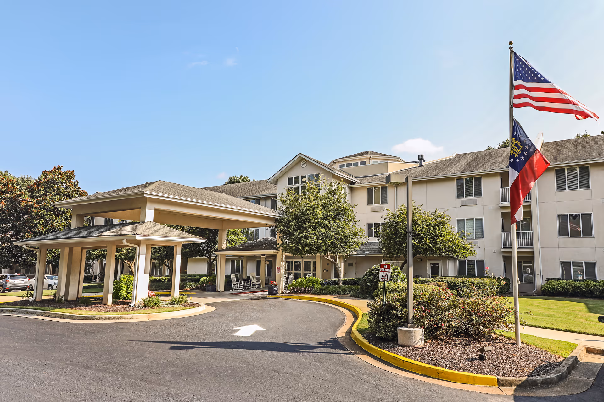 Front entrance of a multi-story senior living building with a covered porte-cochère, landscaped grounds, and flagpoles flying flags.
