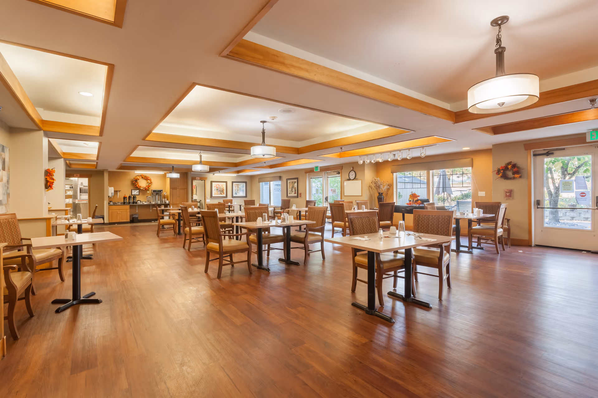 A large, well-lit dining room with multiple tables, chairs, and warm wooden flooring.