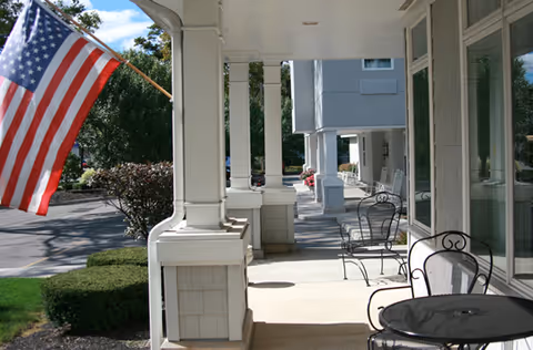 View of a covered outdoor porch area with black metal chairs and tables, white columns, an American flag mounted on the left side, and a parking lot with trees in the background.