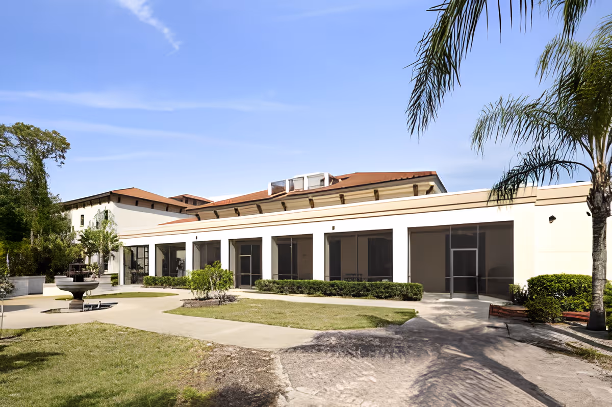 Exterior courtyard and front facade of a single-story building with screened porches, walkways, a fountain, and palm trees.