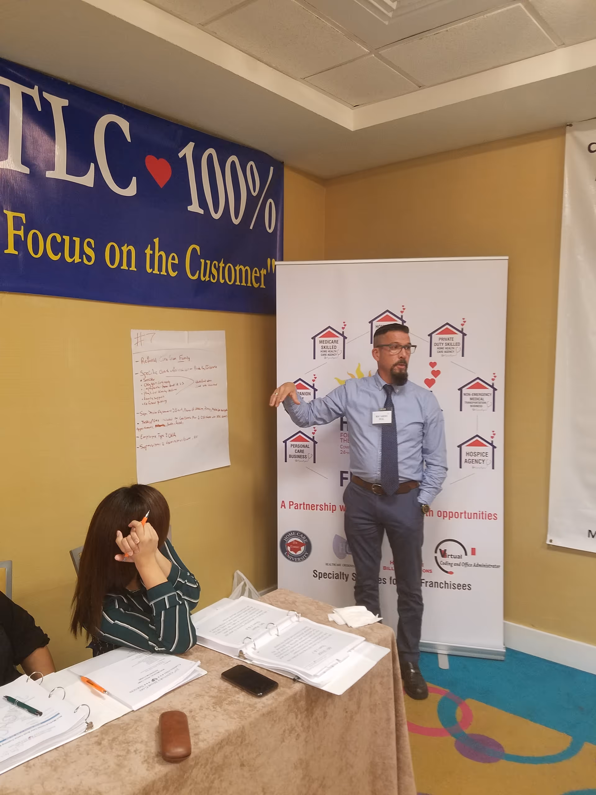 A presenter in a blue shirt speaks beside banners in a meeting room.