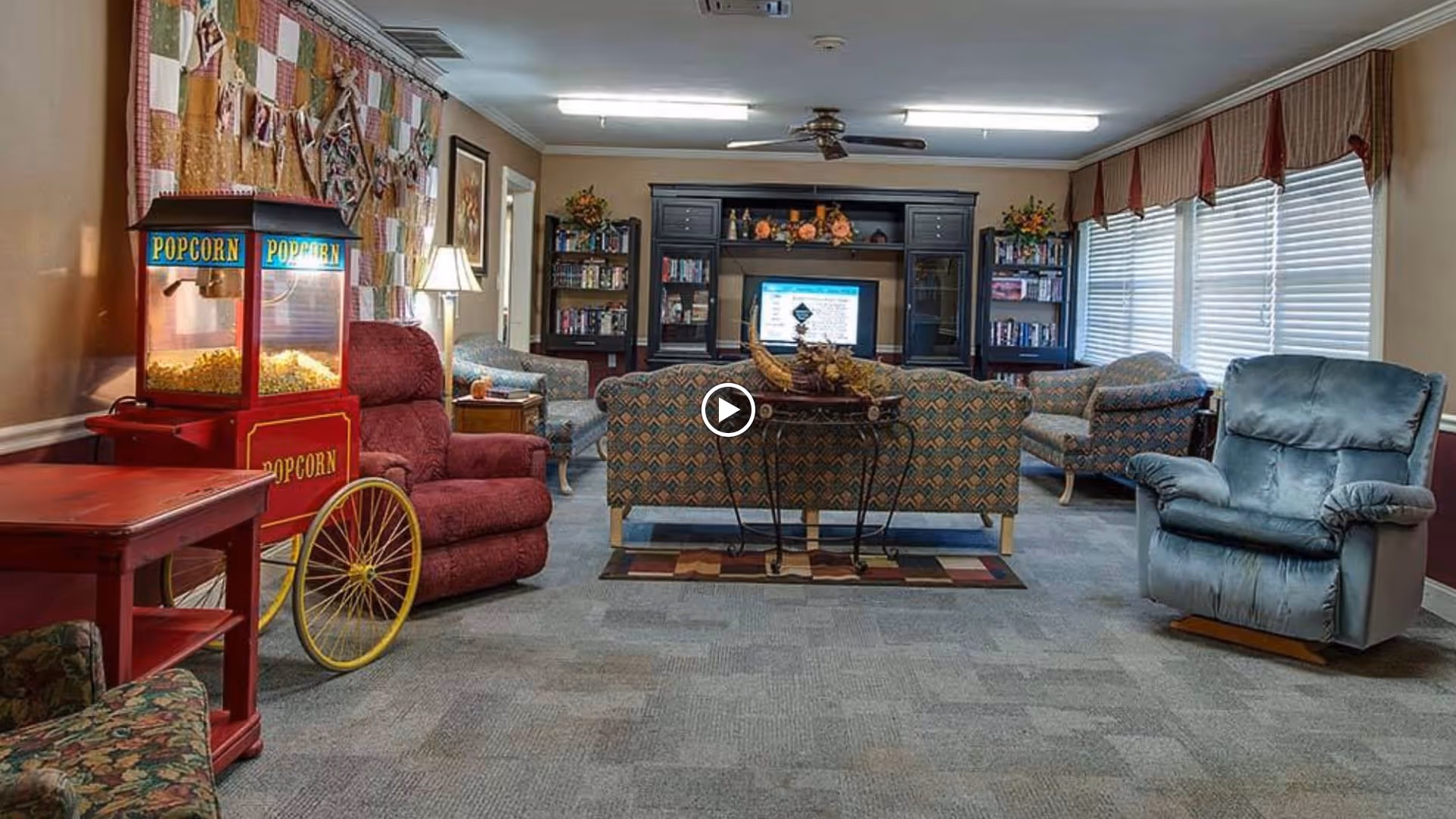 A cozy living room area in a senior living facility with various upholstered chairs and sofas arranged around a TV cabinet. There is a popcorn machine on the left side, a small table with a lamp, bookshelves filled with books, and large windows with blinds and valances on the right side.