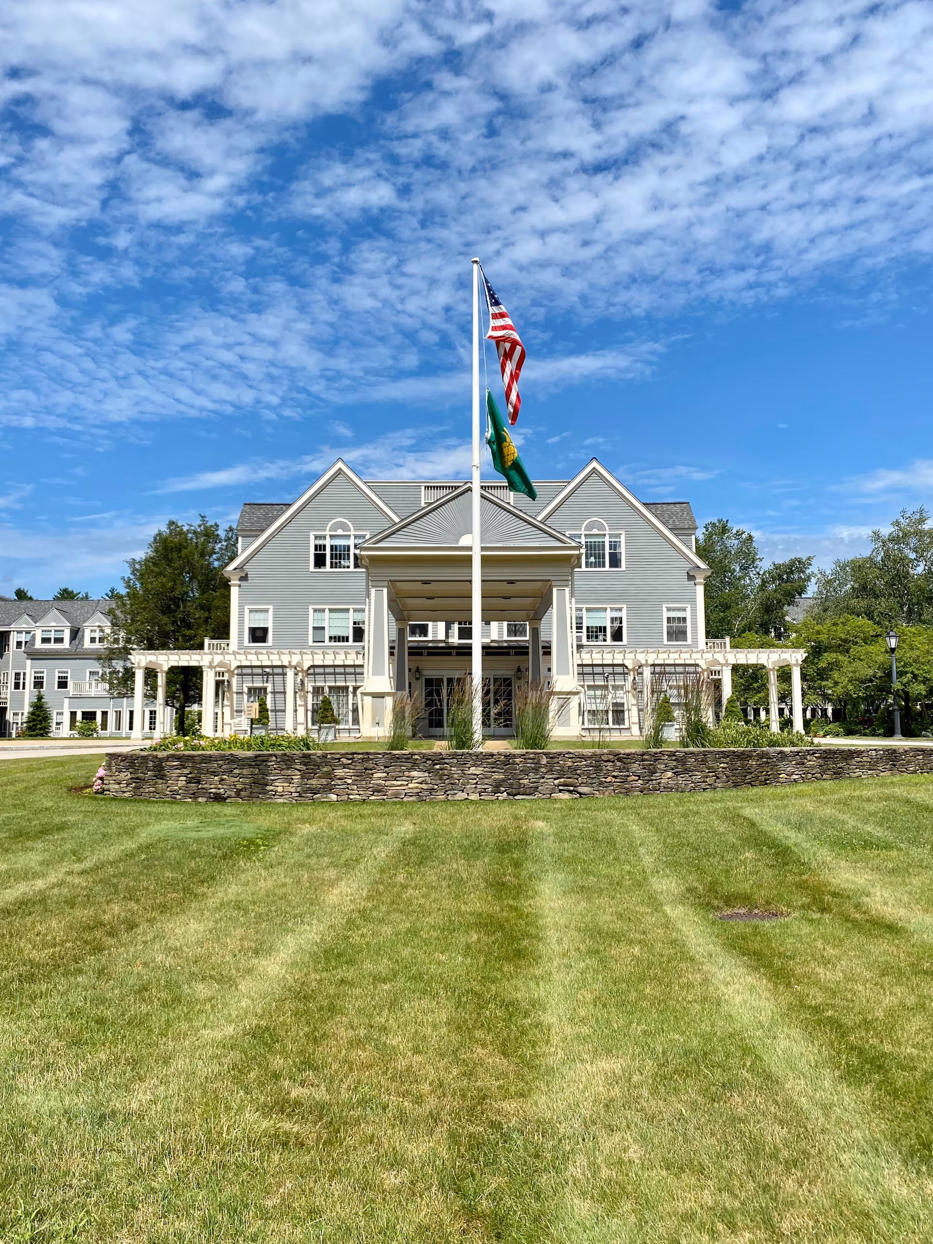 A large senior living facility with a light gray exterior, flagpoles, and a manicured lawn.